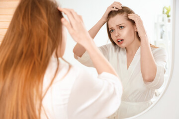Naklejka premium Worried young woman with hair loss problem looking in mirror at home