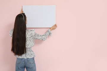 Young woman hanging picture frame on pink wall in room