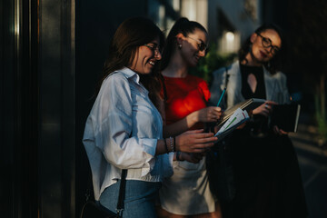 Three businesswomen engaged in a lively discussion outdoors, reviewing project documents and taking notes together.