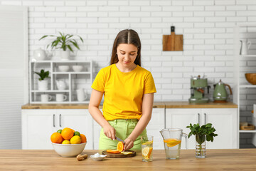 Beautiful young woman cutting orange for preparing lemonade in kitchen