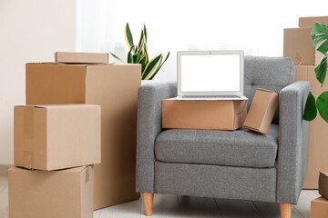 Blank laptop with moving boxes on armchair and plants near window in room