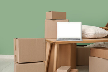 Blank laptop with pillow on table and moving boxes near green wall in room