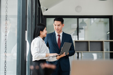 Two business professionals discussing work in a modern office environment, using a laptop and standing near glass walls.