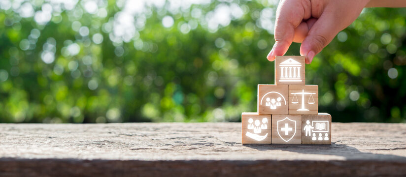 Hand stacking wooden blocks with icons symbolizing social policy elements like healthcare, justice, and community support on a natural background.