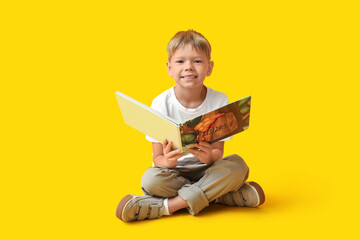 Cute little boy sitting on floor and reading book on yellow background