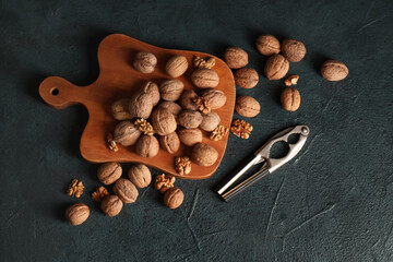 Wooden board with tasty walnuts and nutcracker on dark background