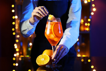 Female bartender decorating glass of Aperol spritz cocktail on table against blurred lights
