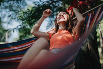 A joyful woman in a red top relaxes in a colorful hammock under a tree, enjoying a sunny summer day.