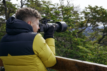 Male photographer in winter jacket captures images with a telephoto lens amidst forest trees.