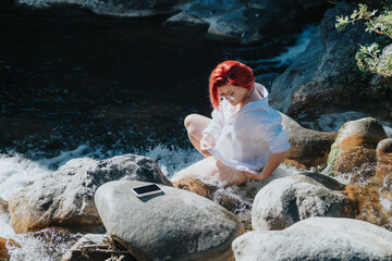 Woman with vibrant red hair wearing a white shirt sitting by a river on rocks, enjoying nature and solitude.