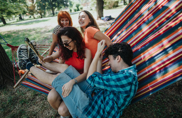 A group of friends laughing and enjoying their time on a colorful hammock in the park on a sunny day.