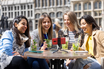 A group of four friends are making memories at an outdoor cafe, taking a selfie while enjoying drinks together