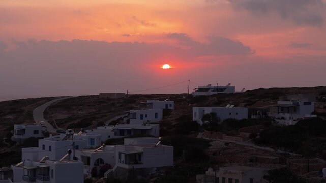 Drone rises above white homes on hillside of Donousa Greece as fire red ball of sun at sunset dips to hide below clouds on ocean