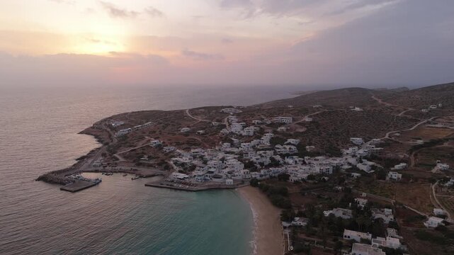 Homes built above port next to beach with clear water at sunset in Donousa Greece, aerial orbit establish