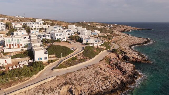 Drone orbits along rocky coastline of Donousa Greece with homes rising along hillside above sandy road