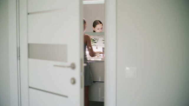 Pretty young girl stands in bathroom near sink, washes hands and wipes them using towel. Adorable teenager stands in front of mirror and makes morning bath routine. Lifestyle. Back view through door.
