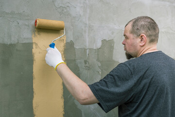 A man in a grey t-shirt paints a vertical strip of tan on a grey wall with a paint roller. The sun...