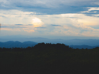 Obraz premium Mountains under a cloudy sky at sunset. Lush greenery in the foreground contrasts beautifully with the misty distant mountains. Serene view of Phu Soi Dao National Park in Thailand.