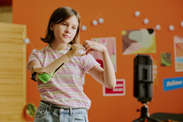Teen girl with arm prosthesis standing in front of smartphone on tripod showing hand heart gesture