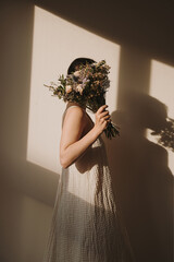 Back view of woman in flowing dress holds a bouquet of flowers, concealing her face. Sunlight casting dramatic shadows indoors, scene with warm, tranquil vibes. Neutral color palette, beige aesthetic