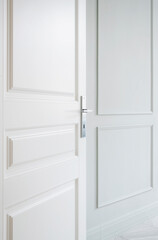 A modern white door stands partially open in a minimalist space featuring soft white walls and elegant paneling. Modern white door partially open in minimalist interior design