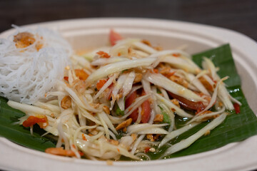 Thai spicy green papaya salad served with rice vermicelli on banana leaf and paper dish. Close up