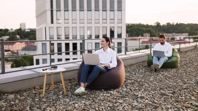 Professionals working on laptops in casual outdoor workspace on rooftop with bean bags, enjoying open air environment. Collaborative work setting reflects modern business and remote work trends.
