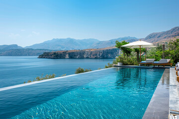 Luxurious Swimming Pool with Blue Water Overlooking the Sea in Kiffos, Crete, Greece, Featuring Lounge Chairs, Umbrella, and Surrounding Greenery with Clear Skies and Distant Mountains