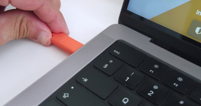 Amazing slow motion close up shot of young man's hand plugging orange lacie disk usb c cable into macbook pro computer in office with natural light and white table.