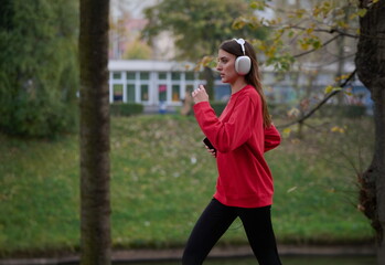 Naklejka premium Young beautiful woman running in autumn park and listening to music with headphones on smartphone