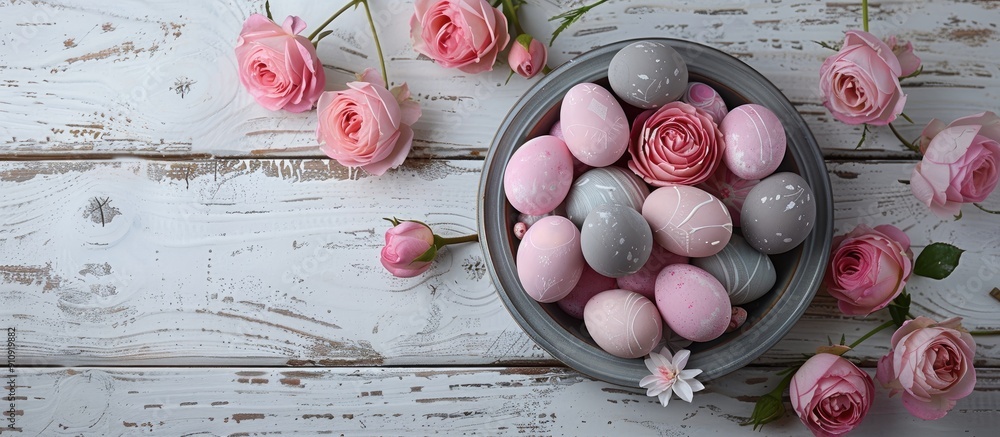 Poster Top view image of Easter eggs in pink and grey hues in a gray dish adorned with roses set on a rustic white wooden table with copy space image