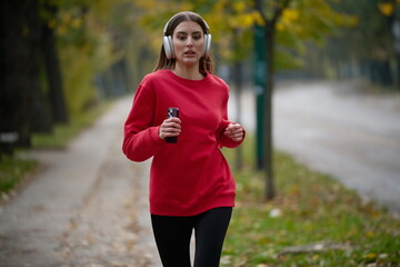 Young beautiful woman running in autumn park and listening to music with headphones on smartphone