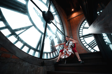A woman sits on a staircase in front of a clock. She is playing a cello. The clock is large and has Roman numerals. The woman is wearing a red dress