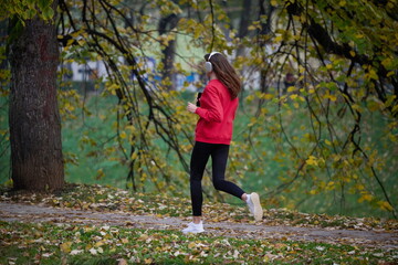 Young beautiful woman running in autumn park and listening to music with headphones on smartphone