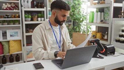Young man working at a laptop in a home decor store, using a scanner while managing paperwork and inventory in a well-decorated interior with shelves of items behind