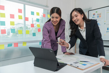 Two Asian businesswomen cheerfully discussing plans in office using calculator and laptop, standing in front of a whiteboard talking.