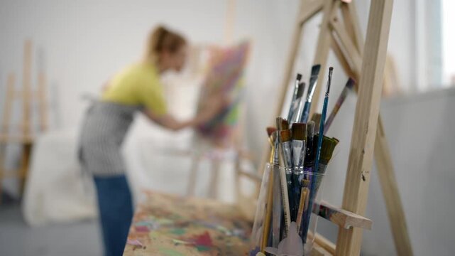 Workshop of professional artist, artistic brushes and lady figure in background. Inspired female painter drawing abstract picture on canvas on easel, work studio of talented woman artist, modern art