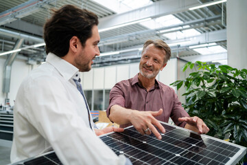 Smiling elderly businessman explaining coworker holding solar panel at factory