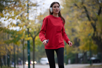 Young beautiful woman running in autumn park and listening to music with headphones on smartphone