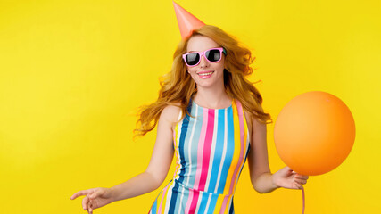 Excited fashionable woman dancing with balloon having fun for celebrate, entertainment. Carefree playful smiling redhead lady, trend makeup, colorful outfit for ad, fashion, party. Studio on yellow