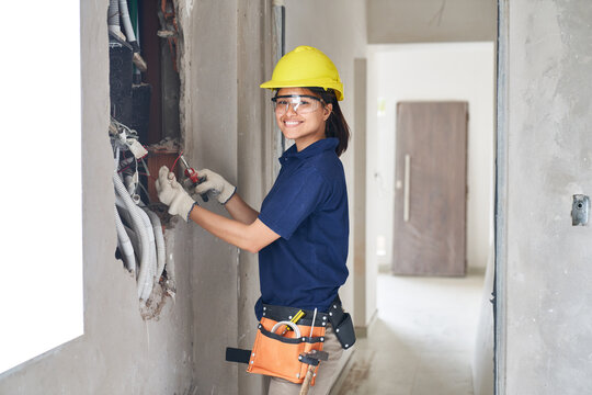 Portrait of smiling young female electrician using pliers while fixing wires in wall at incomplete house construction site