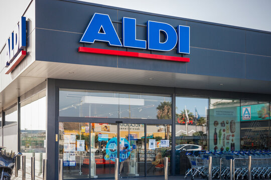 Exterior of an ALDI store. The storefront is visible with clear glass doors and a line of shopping carts, set against a backdrop of a sunny day.
