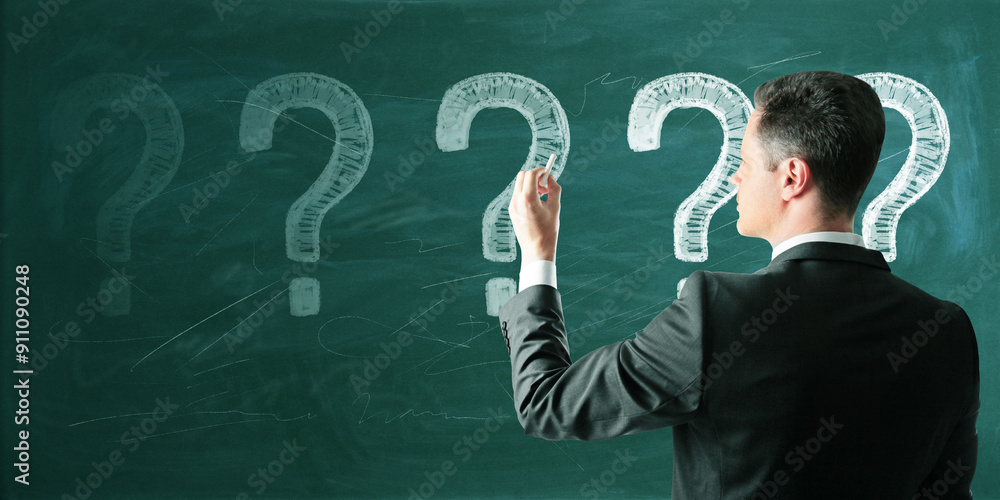 Poster back view of thoughtful young european business man with chalk drawn question marks on blackboard ba
