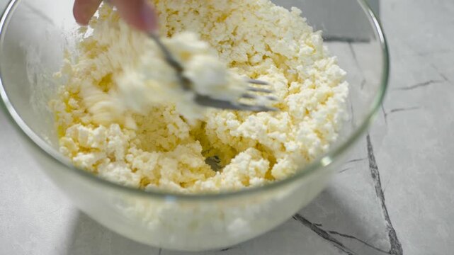 A woman uses a fork to mix cottage cheese with an egg yolk in a transparent bowl. Cooking curd cakes and cheesecakes. Close-up mash and rub cottage cheese with an egg. Pastry