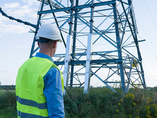 Energy engineer wearing white helmet and reflective vest inspects an electric tower and wires. Energy engineer evaluates power infrastructure, on maintaining and improving electricity distribution.