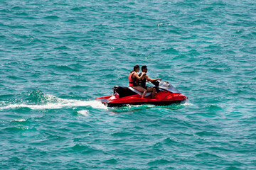 Two people in life jackets on a jet ski speeding through the ocean, creating a wake of splashes.Water transport, summer fun, water sport
