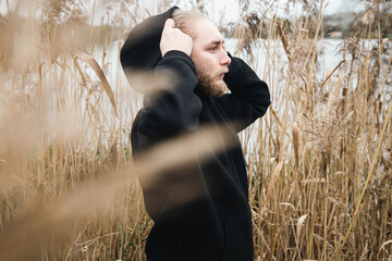 A young bearded man in a black hoodie in the reeds near the river.
