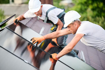 Workers building solar panel system on rooftop of house. Two men installers in helmets installing photovoltaic solar module outdoors. Alternative, green and renewable energy generation concept.