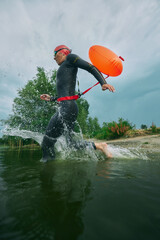 Athlete in wetsuit and swim cap splashes into lake with orange buoy attached, ready for swim amidst...
