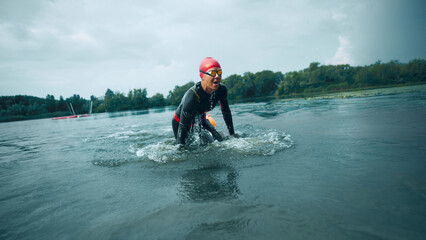 Triathlete in red swim cap and black wetsuit emerges from water, splashing as she transition from...